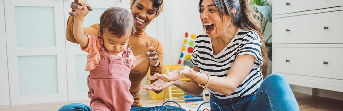 Two women smile as they help a baby take steps. 